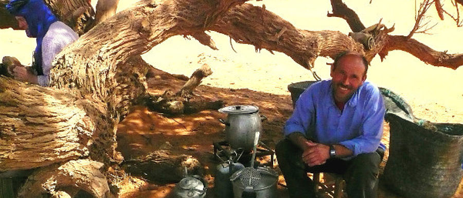 Ein Mann in Hemd und Hose sitzt im Freien auf einem Hocker auf sandigem Boden neben einem Baum. Neben ihm stehen Kochuntensilien. Er schaut in die Kamera. © Robin Menges